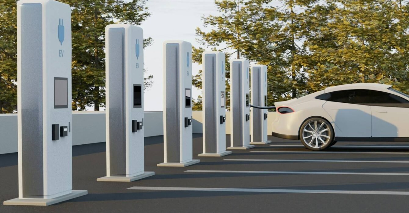 Saudi inter-city EV charging network: A white electric vehicle is charging at a row of EV charging stations amidst trees and a clear sky.